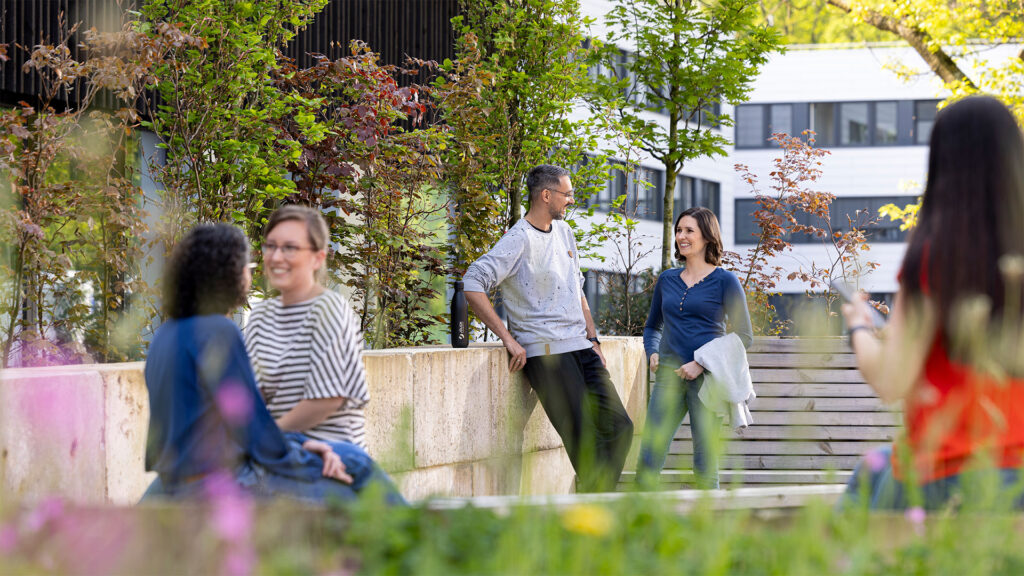 Eine Gruppe von vier Erwachsenen, die sich unterhalten und auf einem sonnigen Platz im Freien mit Bäumen und modernen Gebäuden stehen.