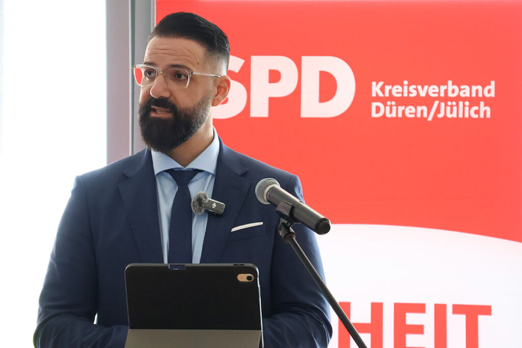 Ein Mann in einem blauen Anzug hält mit einem Mikrofon eine Rede am Rednerpult. Hinter ihm hängt ein rotes Transparent mit der Aufschrift „SPD Kreisverband Düren/Jülich“.
