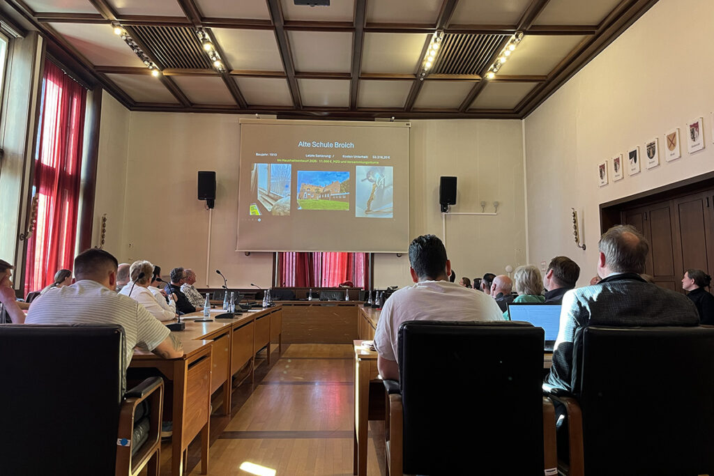 Die Teilnehmer sitzen in einem Besprechungsraum um einen großen Konferenztisch herum und verfolgen eine Präsentation, die auf eine Leinwand an der Vorderseite projiziert wird und Bilder sowie Text enthält.
