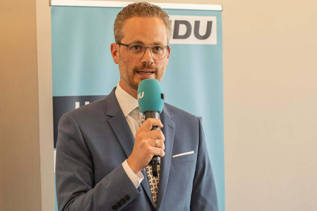 Ein Mann in einem blauen Anzug und mit Brille steht vor einem CDU-Transparent und hält ein türkisfarbenes Mikrofon in der Hand.