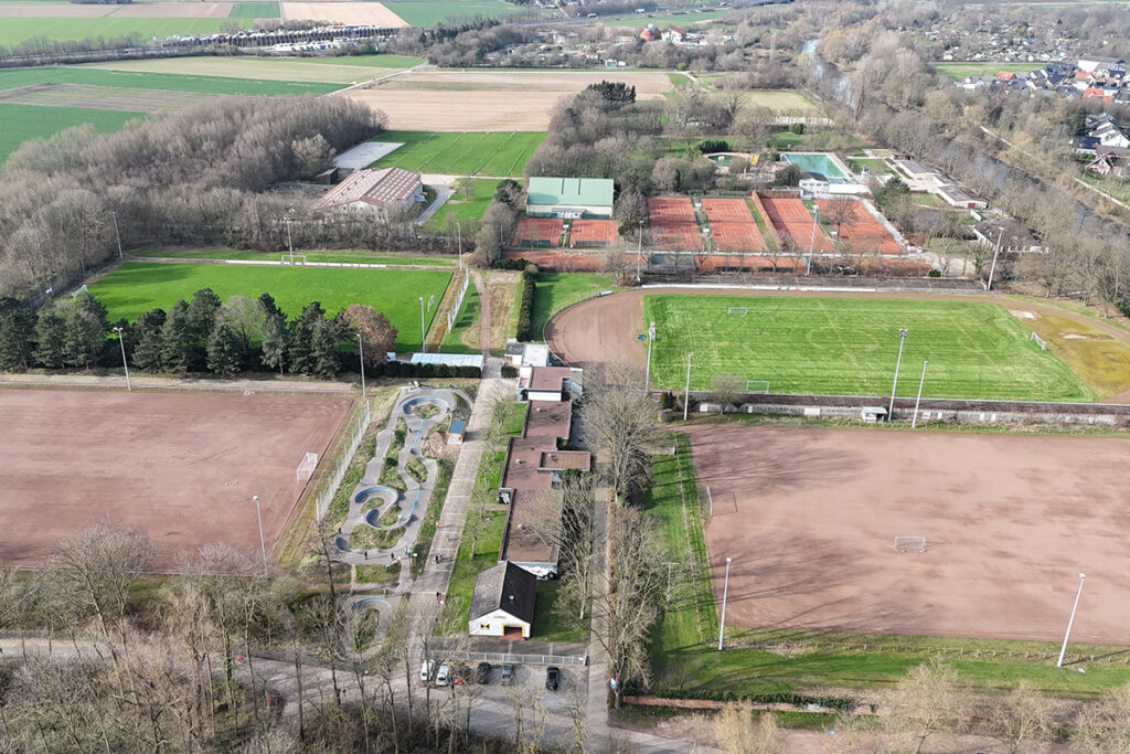Luftaufnahme eines Sportkomplexes mit Tennisplätzen, Laufbahnen, Fußballplätzen, einer BMX-Strecke sowie umliegenden Bäumen und Gebäuden.
