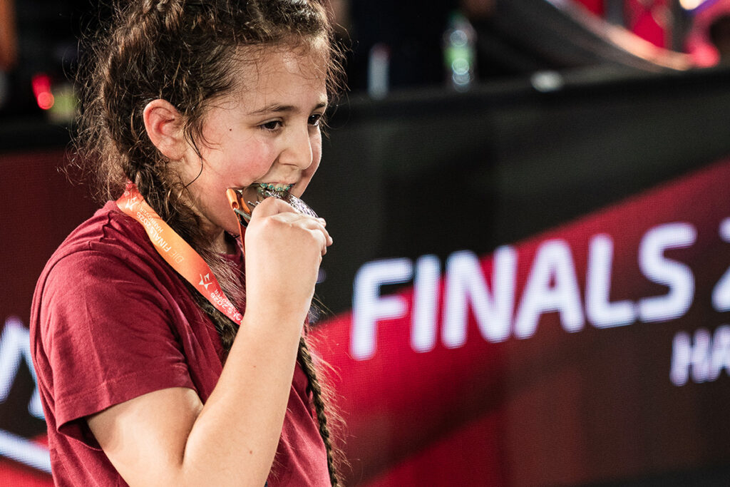Ein junges Mädchen in einem roten Shirt beißt auf eine Medaille, die sie um den Hals trägt, und steht vor einem Schild mit der Aufschrift „FINALS“.