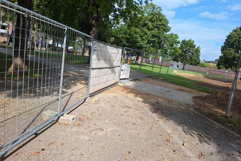 Ein Metallzaun versperrt einen Weg durch einen Park; hinter der Absperrung sind einige Pflastersteine und Baumaterialien zu sehen. Im Hintergrund sind Bäume und Gras unter einem teilweise bewölkten Himmel zu sehen.