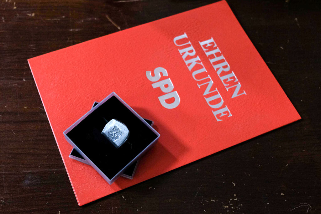 Auf einer dunklen Holzfläche liegt eine Schachtel mit einem silbernen Ring darauf, die auf einer orangefarbenen Urkundenmappe mit der Aufschrift „Ehrenurkunde SPD“ steht.