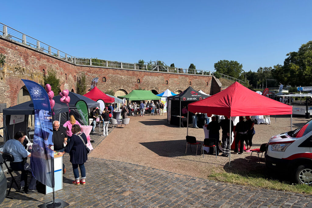 Eine Messe im Freien mit zahlreichen Zelten und Menschen, die an den Ständen miteinander ins Gespräch kommen, vor dem Hintergrund einer Backsteinmauer an einem sonnigen Tag.