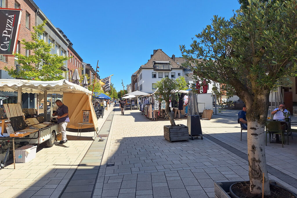 Ein Markt im Freien mit Verkaufsständen, Bäumen in Pflanzkübeln, Menschen, die stöbern und sitzen, sowie Geschäften, die eine sonnige Fußgängerzone säumen.
