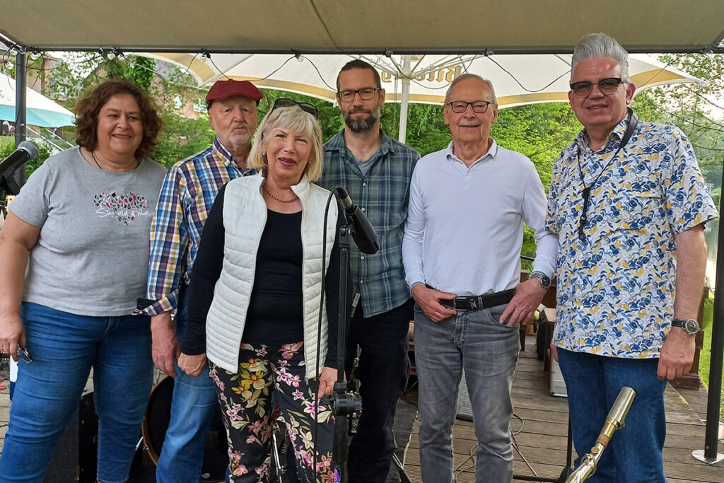 Sechs Erwachsene stehen gemeinsam im Freien unter einem Vordach und posieren für ein Gruppenfoto, wobei im Vordergrund Musikinstrumente zu sehen sind.