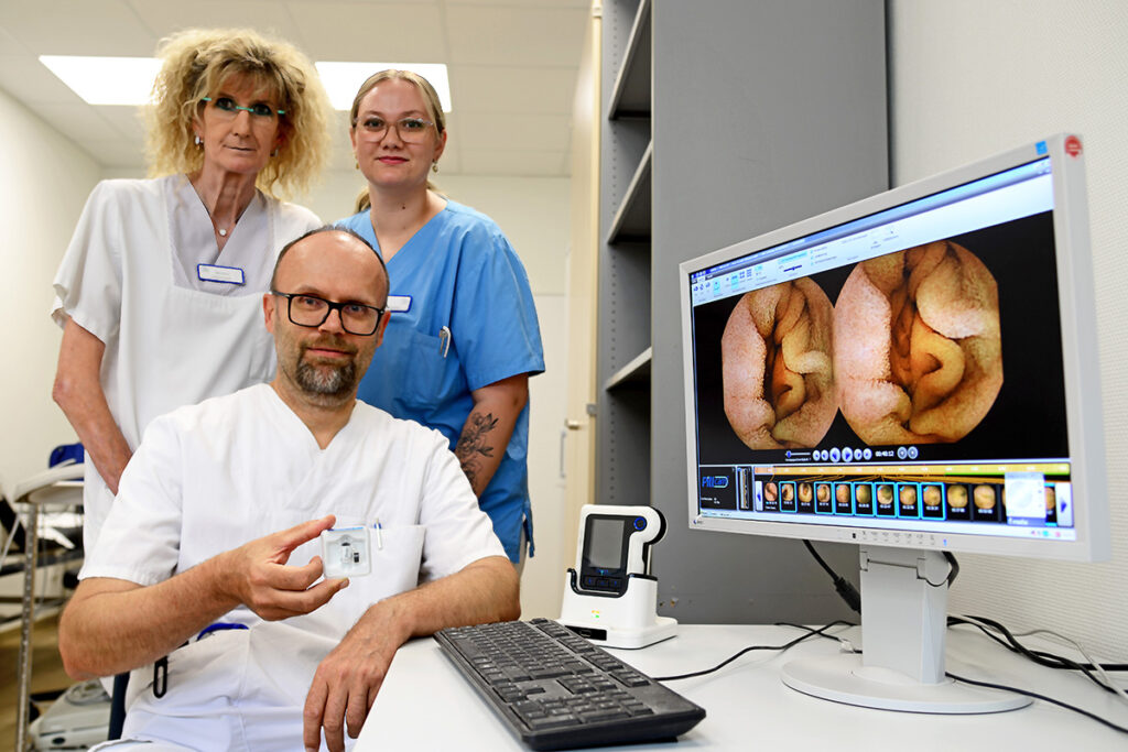 Drei medizinische Fachkräfte posieren vor einem Computer, auf dessen Bildschirm Bilder von inneren Organen zu sehen sind; eine von ihnen sitzt und hält ein medizinisches Gerät in der Hand.