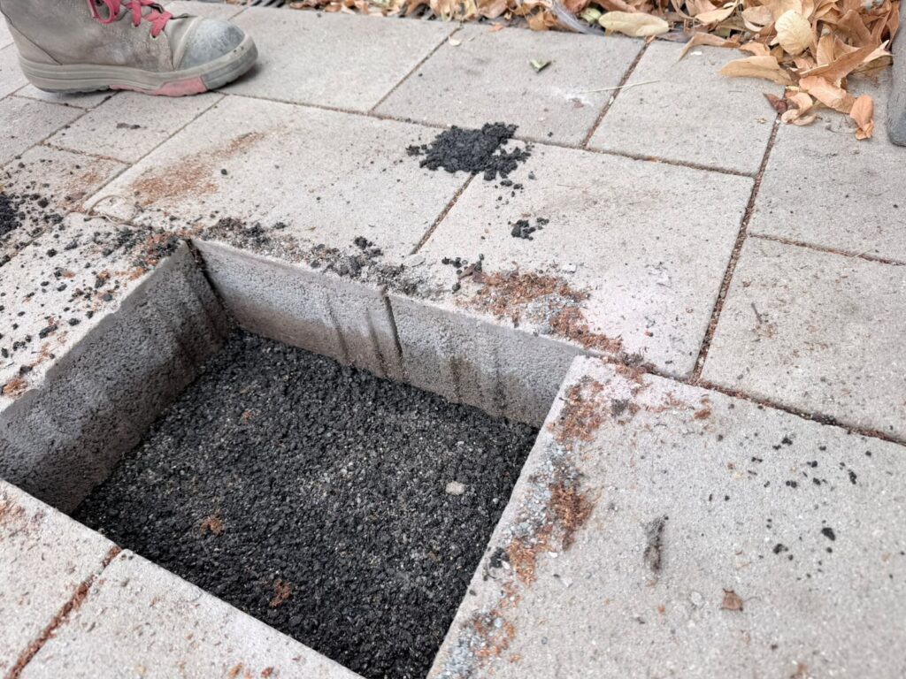 In eine gepflasterte Fläche wurde ein quadratisches Loch geschnitten, wodurch der darunterliegende Kies zum Vorschein kommt. Daneben steht eine Person, die Stiefel trägt. Am oberen Bildrand sind braune Blätter verstreut.
