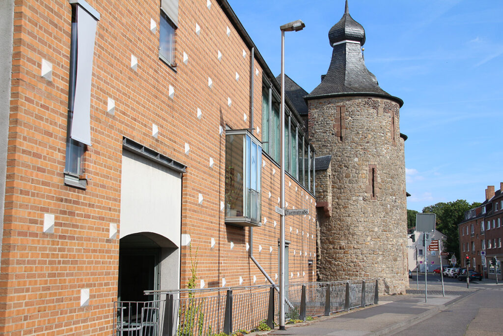 An einer ruhigen Stadtstraße steht unter strahlend blauem Himmel ein Gebäude aus Ziegeln und Stein mit einem Rundturm im mittelalterlichen Stil.