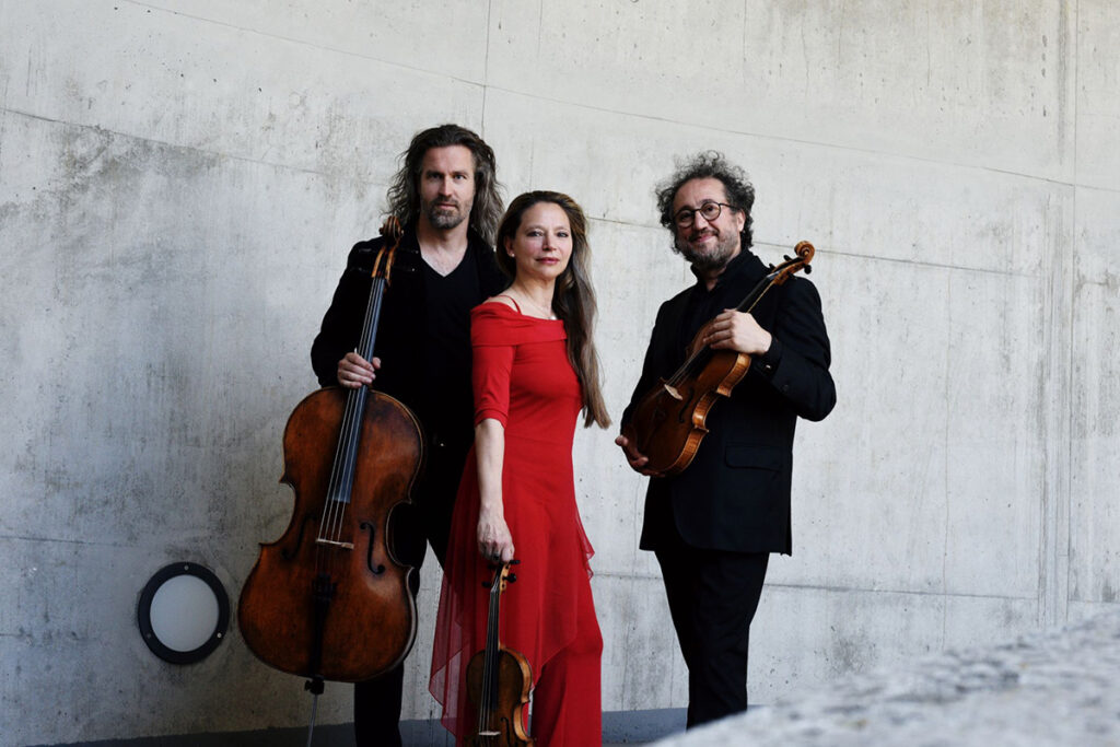Drei Musiker posieren in einem Raum vor einer Betonwand; ein Mann hält ein Cello in der Hand, eine Frau in einem roten Kleid hält eine Geige und ein weiterer Mann hält eine Bratsche.