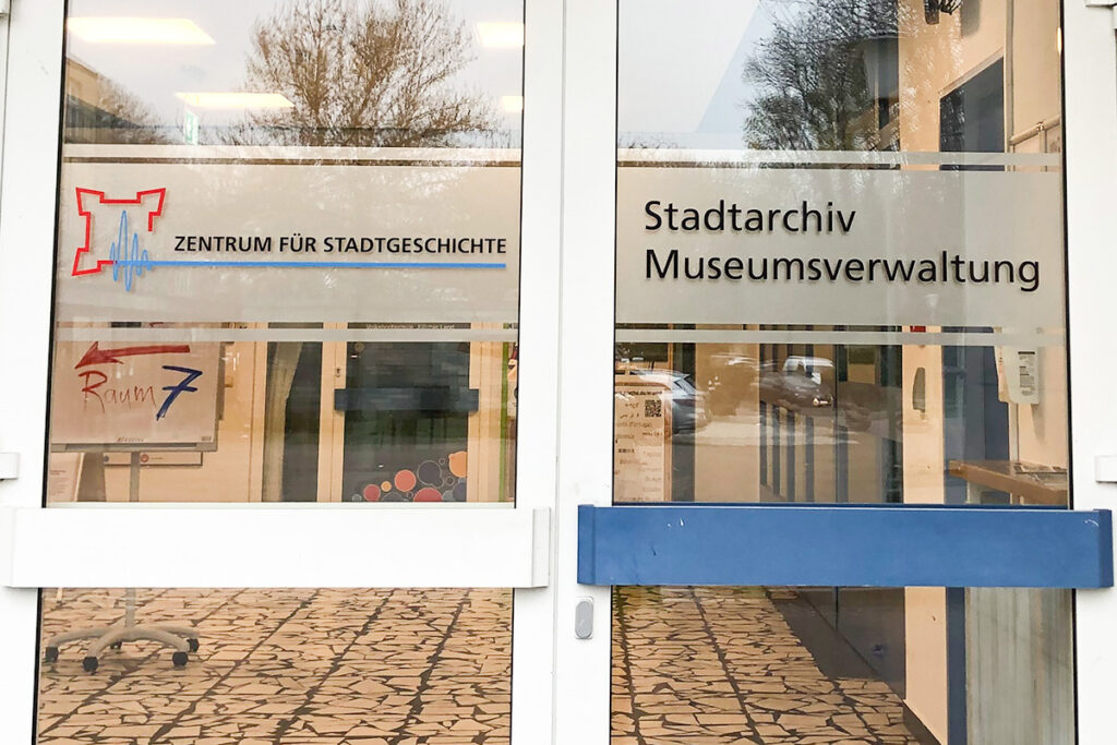 Glastüren mit Schildern für das Zentrum für Stadtgeschichte, das Stadtarchiv und die Museumsverwaltung auf Deutsch, mit Blick auf Bäume und einen Fliesenboden, die sich im Glas spiegeln.