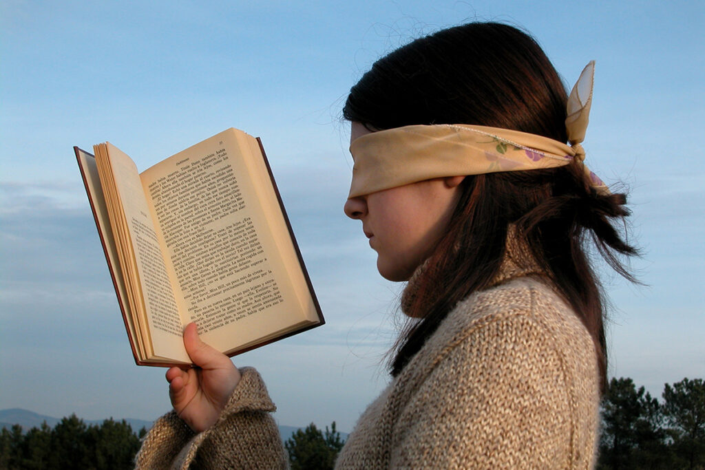 Eine Person mit dunklen Haaren, die einen beigen Pullover und eine Augenbinde trägt, hält im Freien ein aufgeschlagenes Buch in der Hand, im Hintergrund sind Bäume und ein bewölkter Himmel zu sehen.
