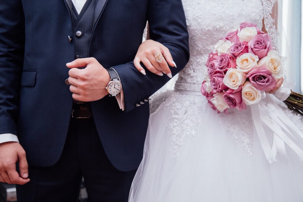 Eine Braut in einem weißen Kleid, die einen Strauß aus rosa und weißen Rosen in der Hand hält, steht Arm in Arm mit einem Bräutigam in einem dunklen Anzug.