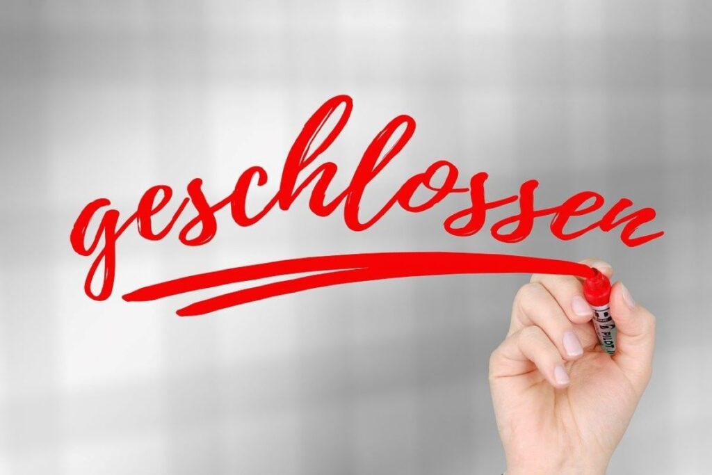 Eine Hand schreibt das deutsche Wort „geschlossen“ mit einem roten Filzstift auf eine transparente Fläche vor einem verschwommenen grauen Hintergrund.