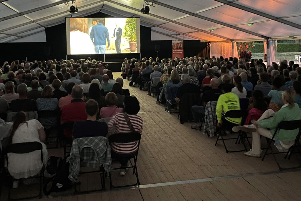 Eine große Gruppe von Menschen sitzt auf Klappstühlen unter einem Zelt und schaut sich einen Film an, der auf eine Leinwand im vorderen Bereich projiziert wird.