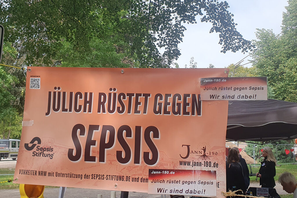 Auf einem orangefarbenen Banner steht „Jülich rüstet gegen Sepsis“ – im Hintergrund einer Freiluftveranstaltung sind Bäume und Menschen zu sehen. Kleinere Schilder greifen die Botschaft zur Aufklärung über Sepsis und zur Unterstützung auf.