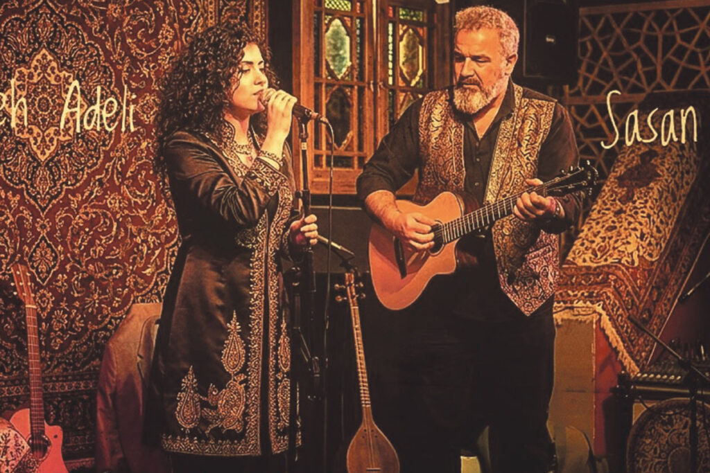 Eine Frau singt ins Mikrofon, während ein Mann neben ihr Gitarre spielt. Beide tragen bestickte Kleidung und stehen auf einer mit Teppich ausgelegten Bühne, im Hintergrund sind gemusterte Teppiche zu sehen.