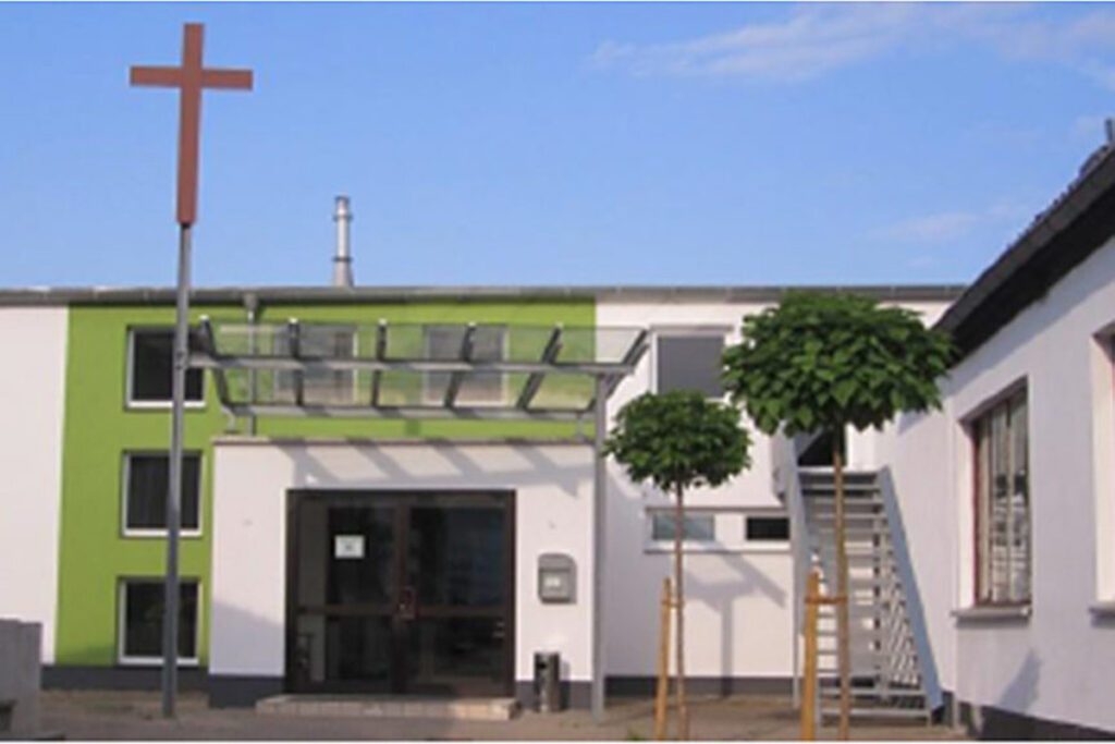 Ein modernes Kirchengebäude mit einem großen Kreuz, grün-weißen Wänden, einem verglasten Eingangsbereich, zwei kleinen Bäumen und einer Außentreppe.