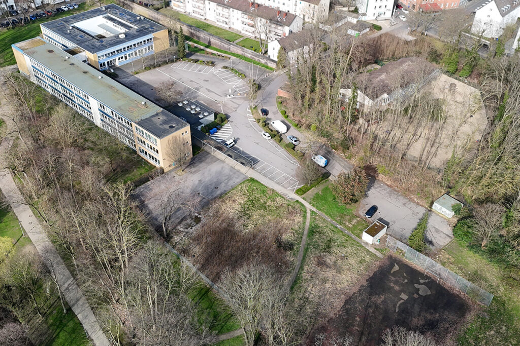 Luftaufnahme eines rechteckigen Gebäudes mit einem Parkplatz, mehreren geparkten Autos, angrenzenden Bäumen und nahegelegenen Wohngebäuden.