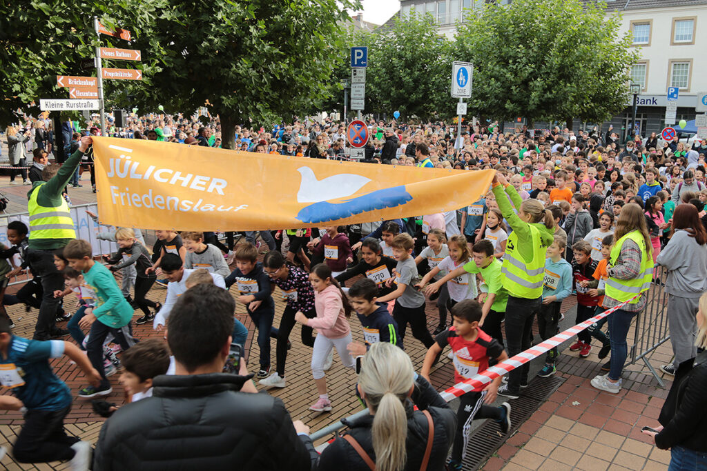 Eine große Gruppe von Kindern nimmt an einem Straßenlauf unter einem orangefarbenen Banner mit der Aufschrift Jülicher Friedenslauf teil, umgeben von Zuschauern und Veranstaltungsmitarbeitern in gelben Westen.