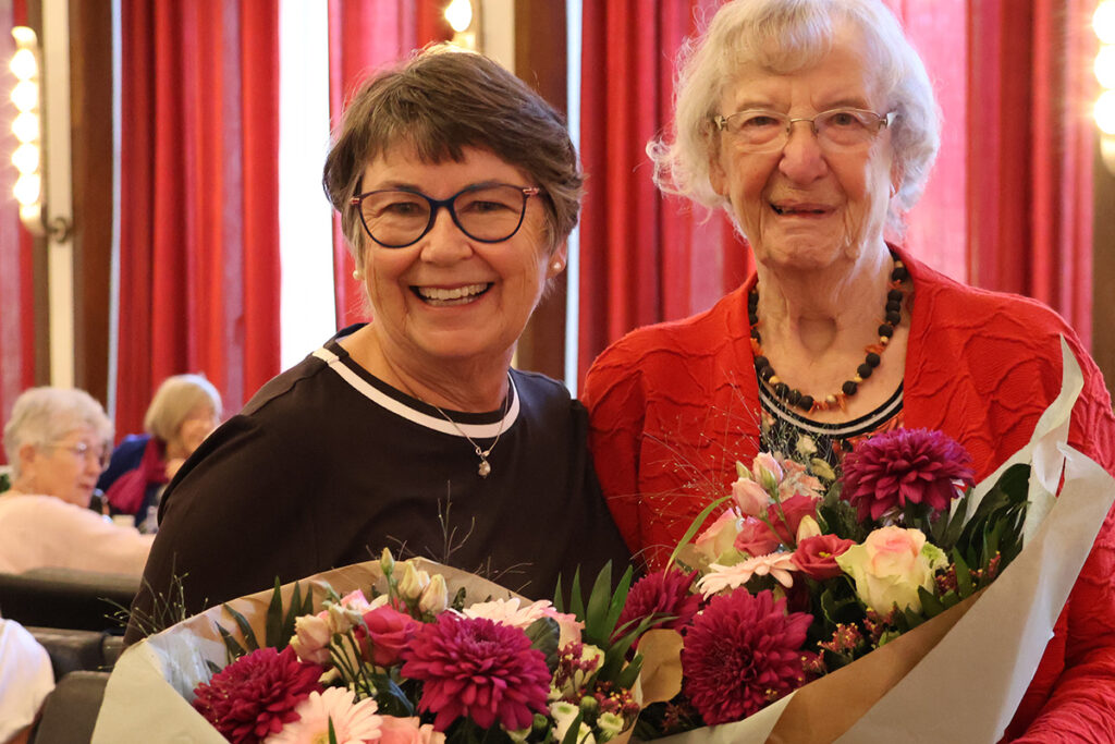 Zwei Frauen stehen in einem Raum, lächeln und halten Blumensträuße in den Händen. Im Hintergrund sind rote Vorhänge und weitere sitzende Personen zu sehen.