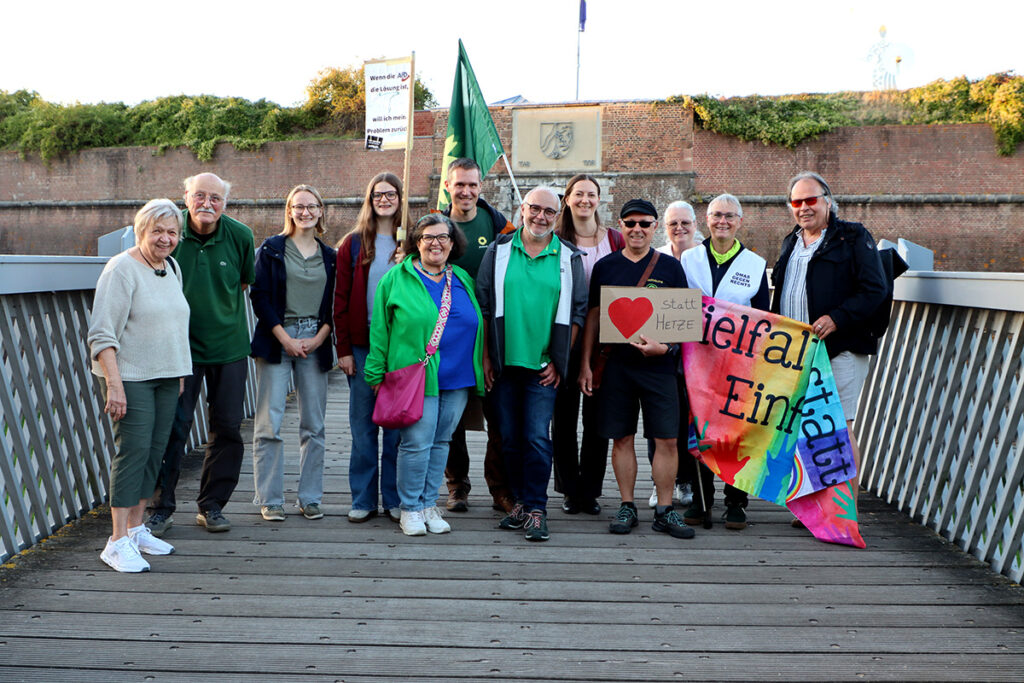 Eine Gruppe von Menschen posiert auf einer Brücke und hält ein buntes Transparent sowie ein Schild mit der Aufschrift „Herz statt Hetze“ in den Händen. Einige lächeln und halten grüne Fahnen in den Händen. Im Hintergrund sind eine Backsteinmauer und Grünflächen zu sehen.