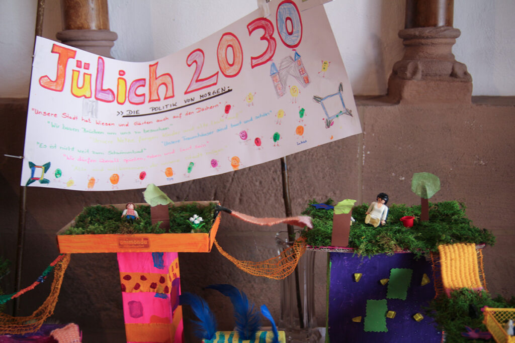 Handgefertigtes Diorama mit zwei kleinen Figuren und Pflanzen unter einem bunten „Jülich 2030“-Schild, das im Innenbereich vor einer Steinwand ausgestellt ist.