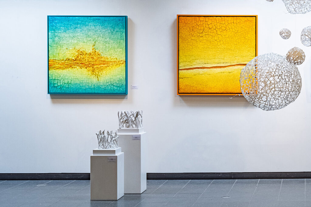 An einer Galeriewand hängen zwei abstrakte Gemälde mit Struktur, eines in Blaugrün und eines in Gelb, über drei kleinen weißen Skulpturen und neben kugelförmigen Drahtkunstwerken.