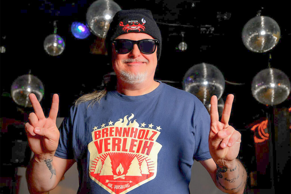 Ein Mann, der eine Sonnenbrille, eine Mütze und ein T-Shirt von „Brennholz Verleih“ trägt, zeigt mit beiden Händen das Peace-Zeichen. Im Hintergrund hängen mehrere Discokugeln.