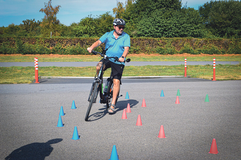 Ein Mann mit Helm und blauem Hemd fährt mit dem Fahrrad durch einen Parcours mit blauen, grünen und roten Kegeln auf einem Bürgersteig im Freien.