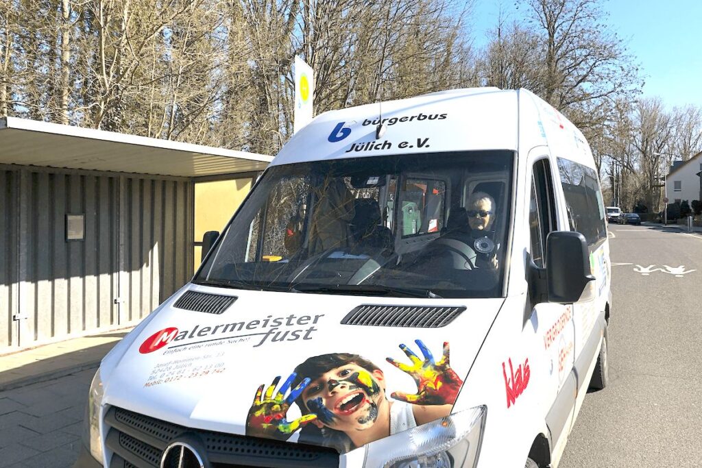 Ein weißer Lieferwagen mit bunter Werbung, darunter das Foto eines Kindes mit bemalten Händen, steht an einem sonnigen Tag an einer Bushaltestelle, im Hintergrund sind Bäume und Gebäude zu sehen.