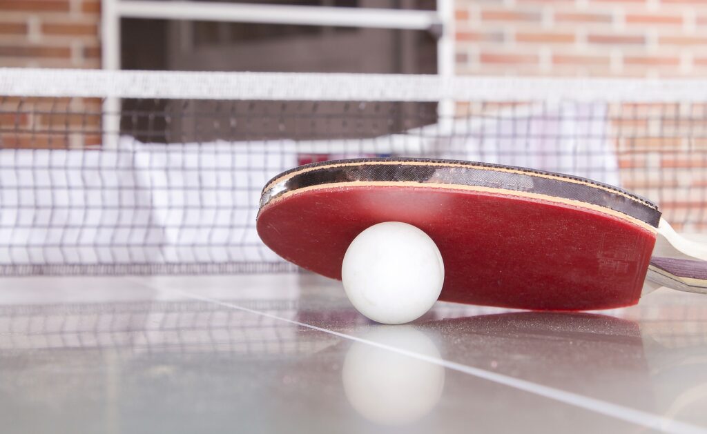 Eine Nahaufnahme eines roten Tischtennisschlägers, der auf einem weißen Ball auf einem Tischtennistisch ruht, im Hintergrund sind ein Netz und eine Backsteinmauer zu sehen.