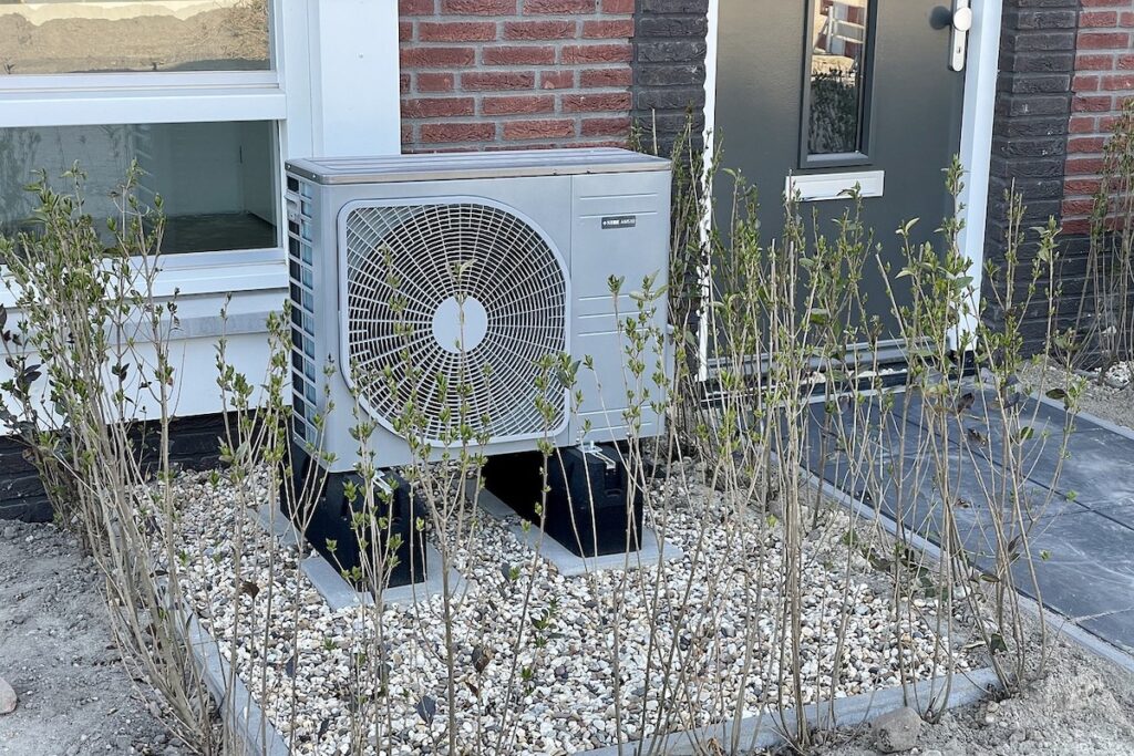 Eine Wärmepumpenanlage ist auf einem Kiesbett mit kleinen grünen Sträuchern vor einem Haus mit Ziegel- und Fassadenverkleidung installiert.