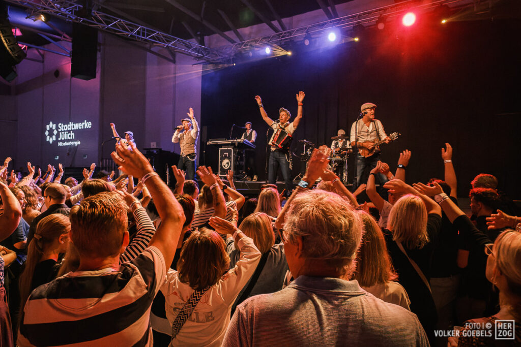Eine Band, die auf der Bühne vor einem jubelnden Publikum auftritt, das die Arme in die Höhe reckt.