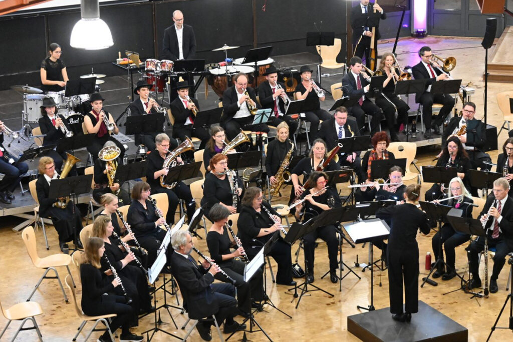 Ein vollbesetztes Orchester, das auf der Bühne auftritt, mit Musikern, die verschiedene Instrumente spielen, und einem Dirigenten, der auf dem Podium steht.