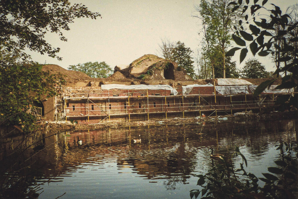 Ein teilweise restauriertes Backsteingebäude mit Gerüsten steht an einem Teich, umgeben von Bäumen und Laub, auf dessen Wasser Enten schwimmen.