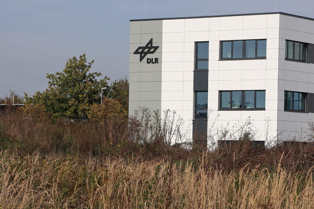 Ein weißes Bürogebäude mit dem DLR-Logo an der Fassade, das hinter einem Feld mit trockenem Gras und Bäumen steht.