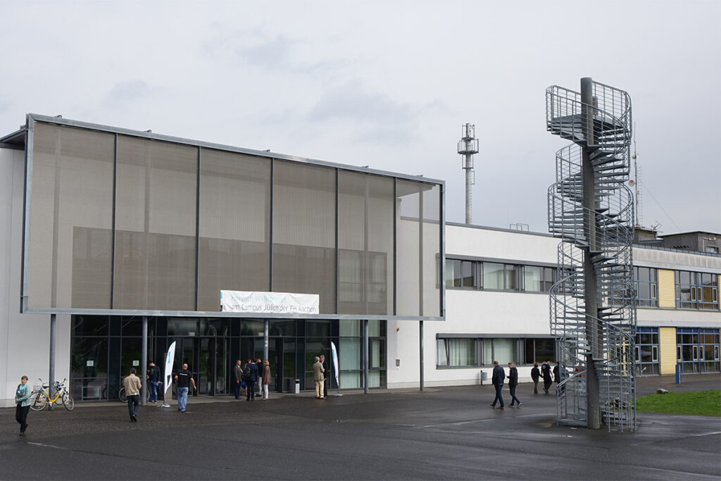 Menschen, die vor dem Campusgebäude der FH Aachen in Jülich vorbeigehen, das sich durch eine Metallgitterfassade und eine markante Wendeltreppe auszeichnet.