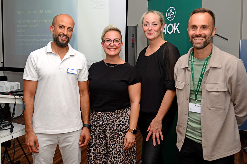 Vier Personen stehen bei einer Veranstaltung in einem Saal in einer Reihe, im Hintergrund sind eine Präsentationsfolie und ein Banner mit dem AOK-Logo zu sehen.