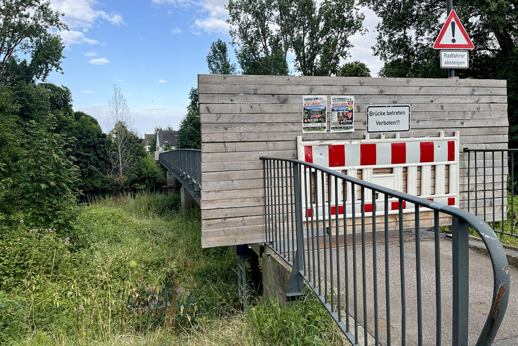 Eine Holzbarrikade versperrt den Zugang zu einer Fußgängerbrücke; darauf stehen Schilder mit der Aufschrift Brücke betreten verboten!!! sowie eine Warnung an Radfahrer, abzusteigen.