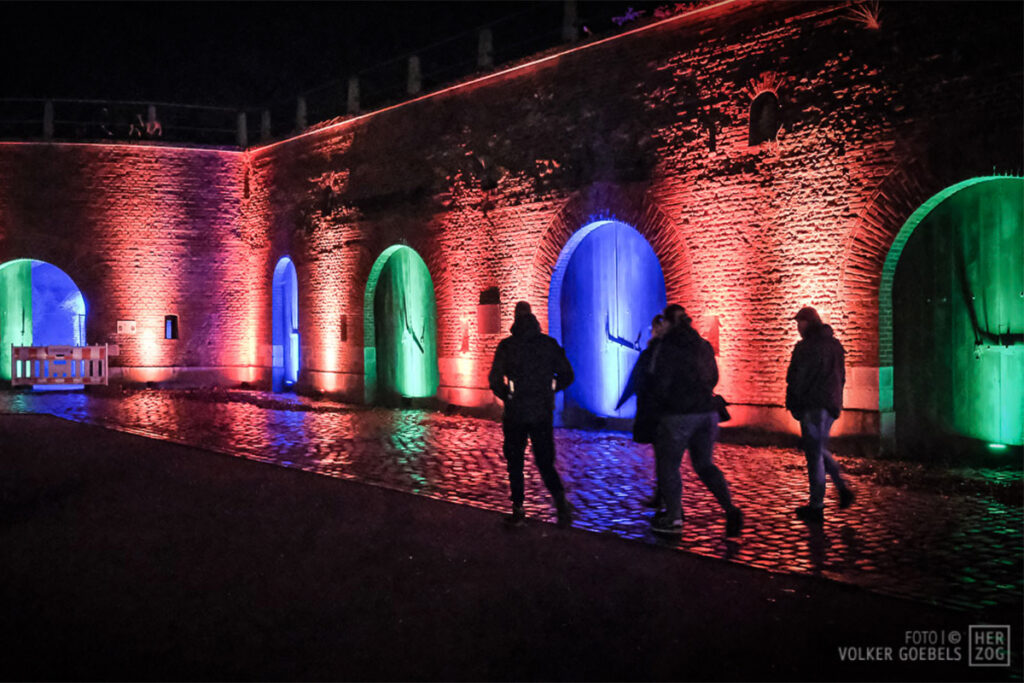 Menschen, die an den mit roten, blauen und grünen Lichtern beleuchteten Backsteinmauern der Festung vorbeigehen.