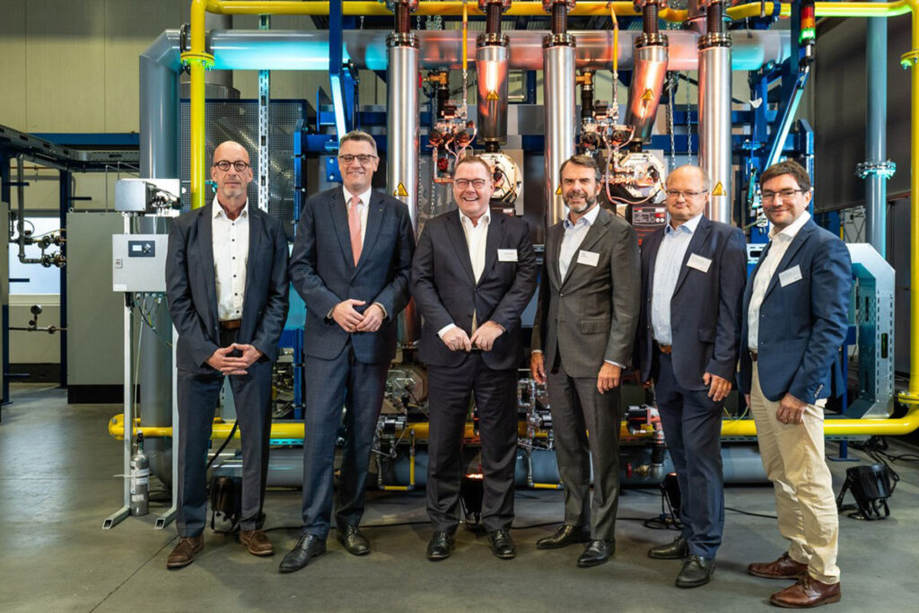 Sechs Männer in Geschäftskleidung, die in einer Reihe vor Industriemaschinen stehen.
