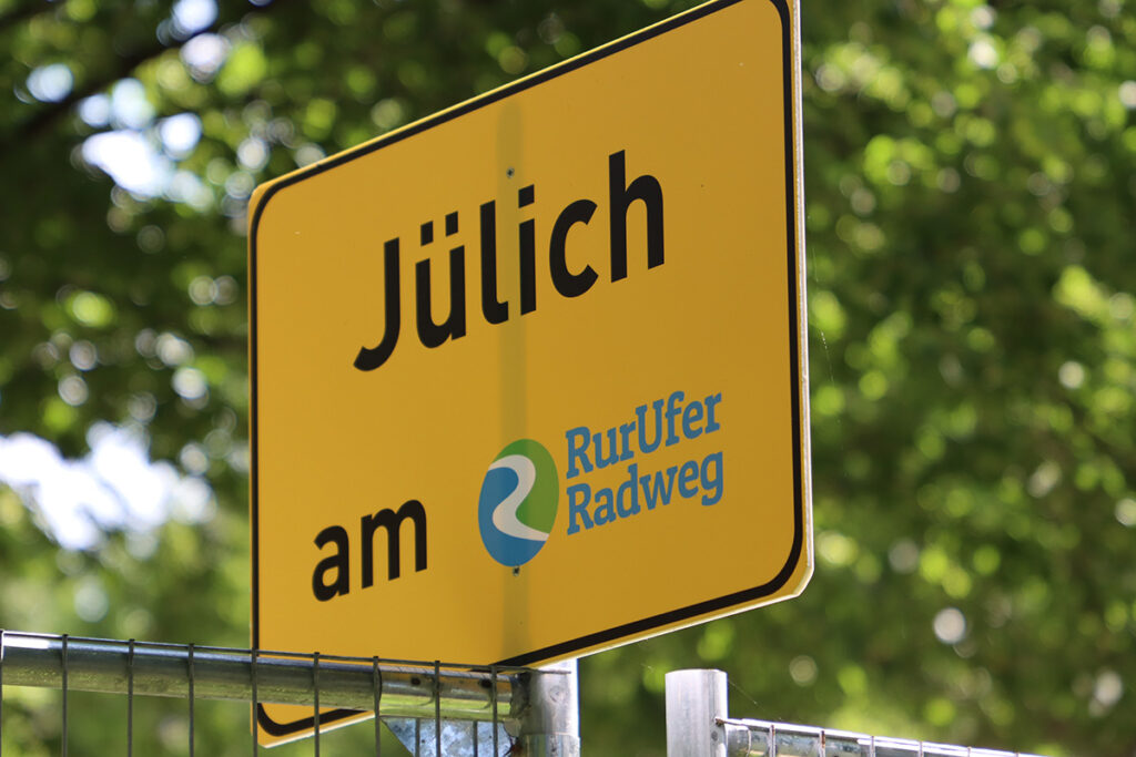 Gelbes Ortsschild mit der Aufschrift Jülich am RurUfer Radweg und einem blau-weißen, runden Logo.