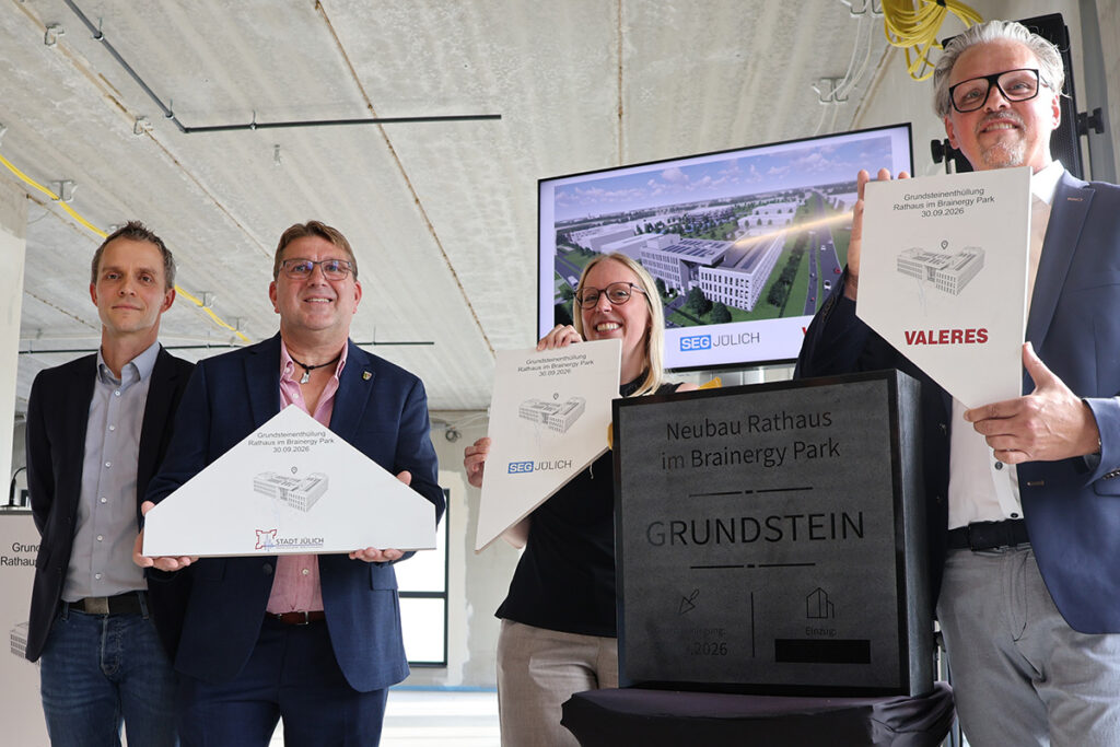 Vier Personen stehen in einem Innenraum neben einem Grundstein und halten Plaketten in den Händen, während im Hintergrund auf einem Bildschirm eine Gebäudevisualisierung zu sehen ist.
