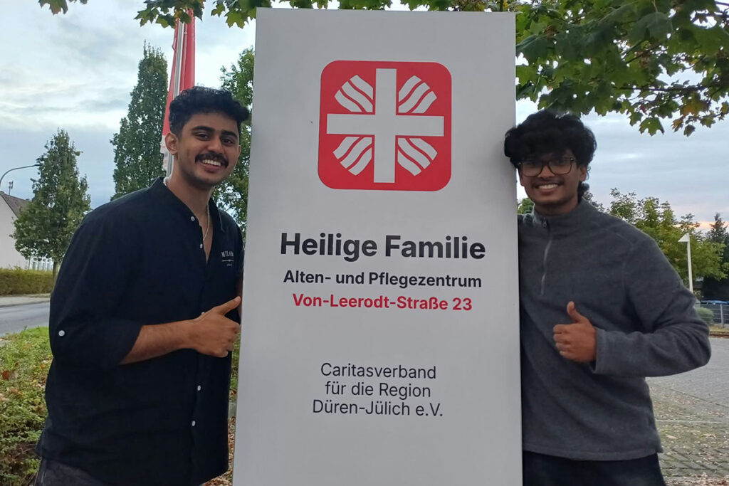 Zwei junge Männer posieren neben einem Schild des Pflegeheims Heilige Familie in Düren-Jülich.
