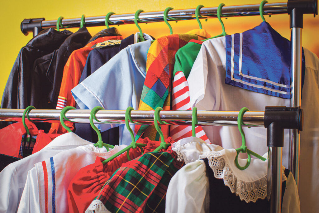Auf einem Kleiderständer hängen verschiedene farbenfrohe Kostüme und Uniformen an grünen Kleiderbügeln, darunter karierte, gestreifte und Outfits im Matrosenstil, vor einem gelben Hintergrund.