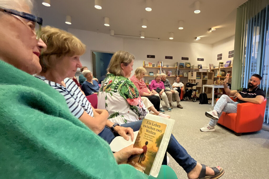 Eine Gruppe von Menschen sitzt in einer Bibliothek, einige halten Bücher in der Hand, während ein Mann in einem roten Sessel im Rahmen einer Diskussion oder einer Lesung zu ihnen spricht.