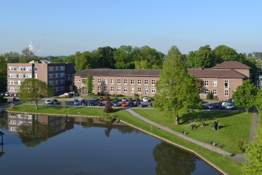 Ein mehrstöckiger Gebäudekomplex aus Backstein mit Parkplätzen, grünen Rasenflächen und einem Teich im Vordergrund.