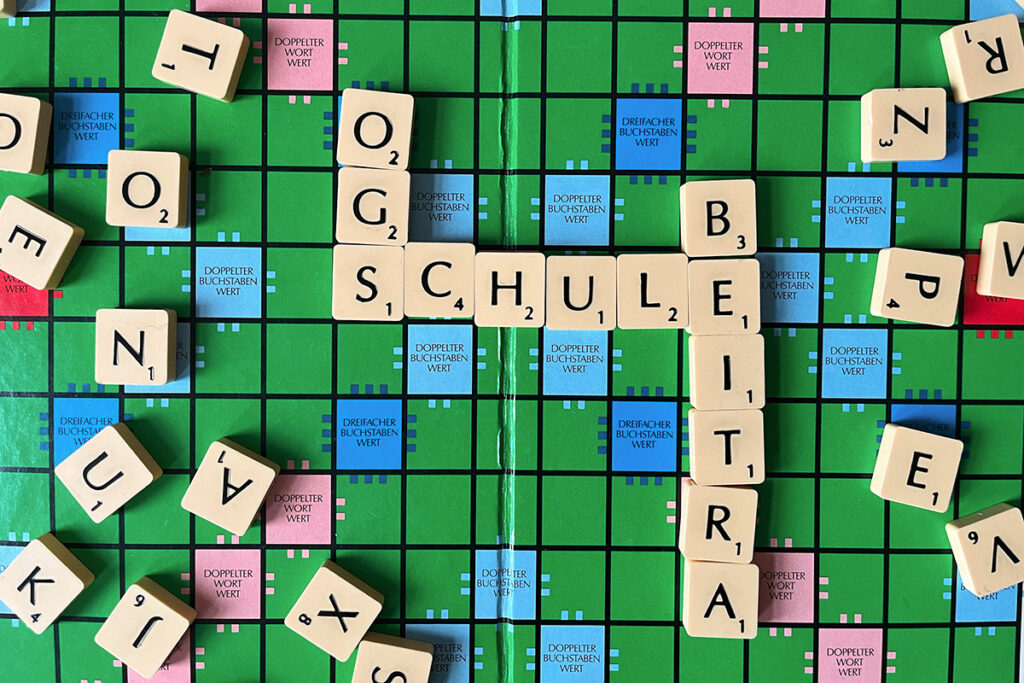 Ein Scrabble-Spielbrett mit dem deutschen Wort SCHULE, das von BEITRAG durchkreuzt wird, wobei lose Buchstabenplättchen am Rand verstreut liegen.